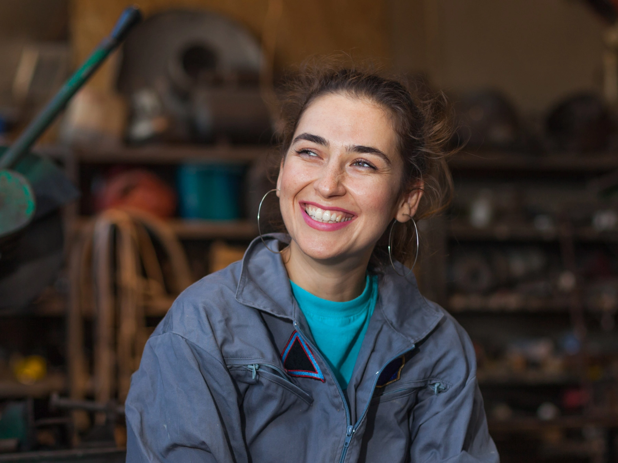 Junge Frau im Blaumann sitzt lächelnd in einer Werkstatt.